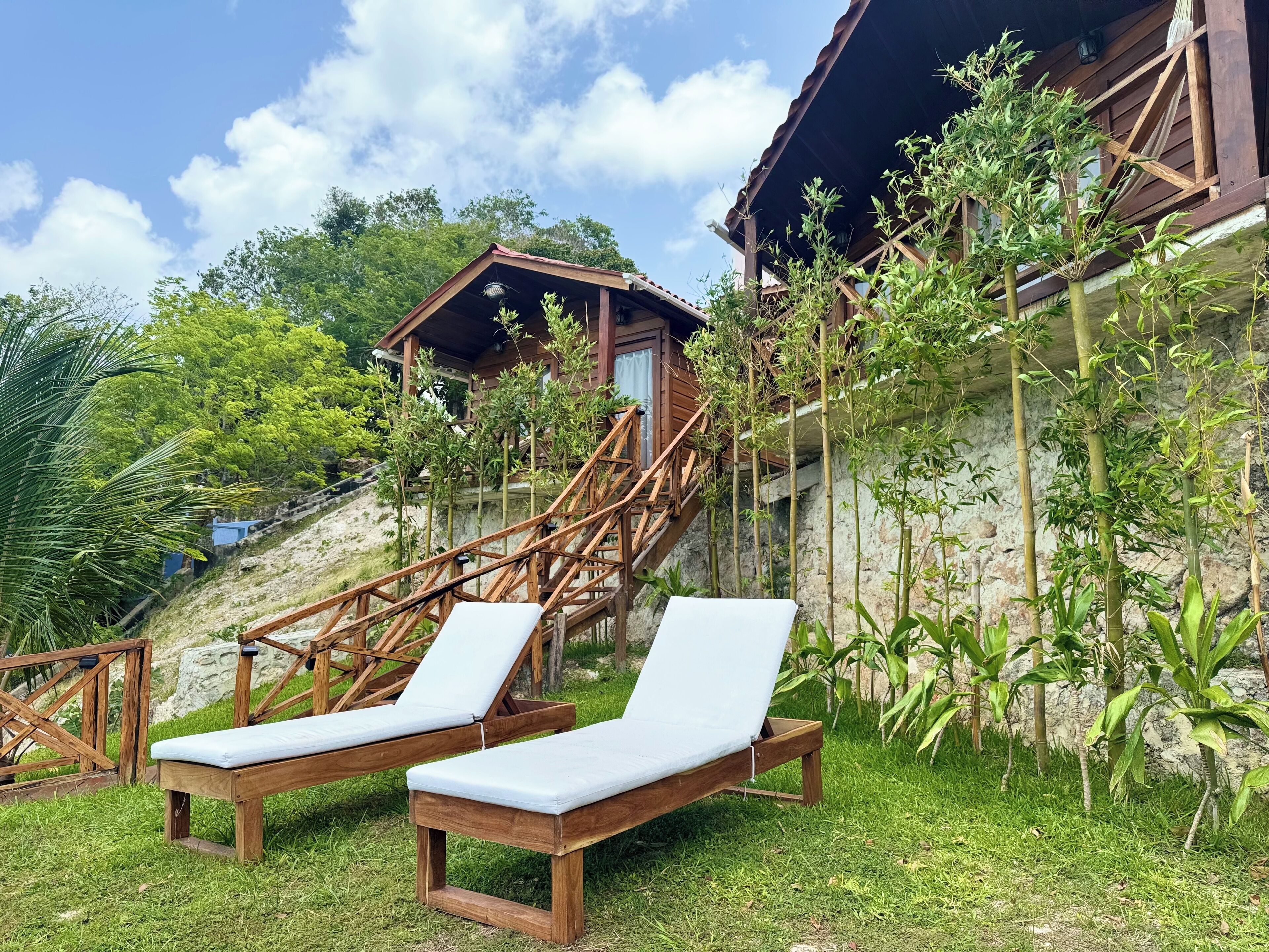 Un espace extérieur aménagé en bois, avec deux fauteuils de jardin, un escalier menant à un bâtiment en bois et une végétation luxuriante.