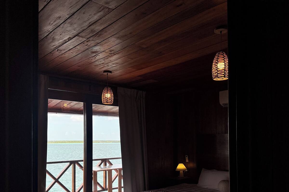 A bedroom with a wooden ceiling, a bed with a white duvet, a nightstand with a lamp, and a view of the sea through large windows.