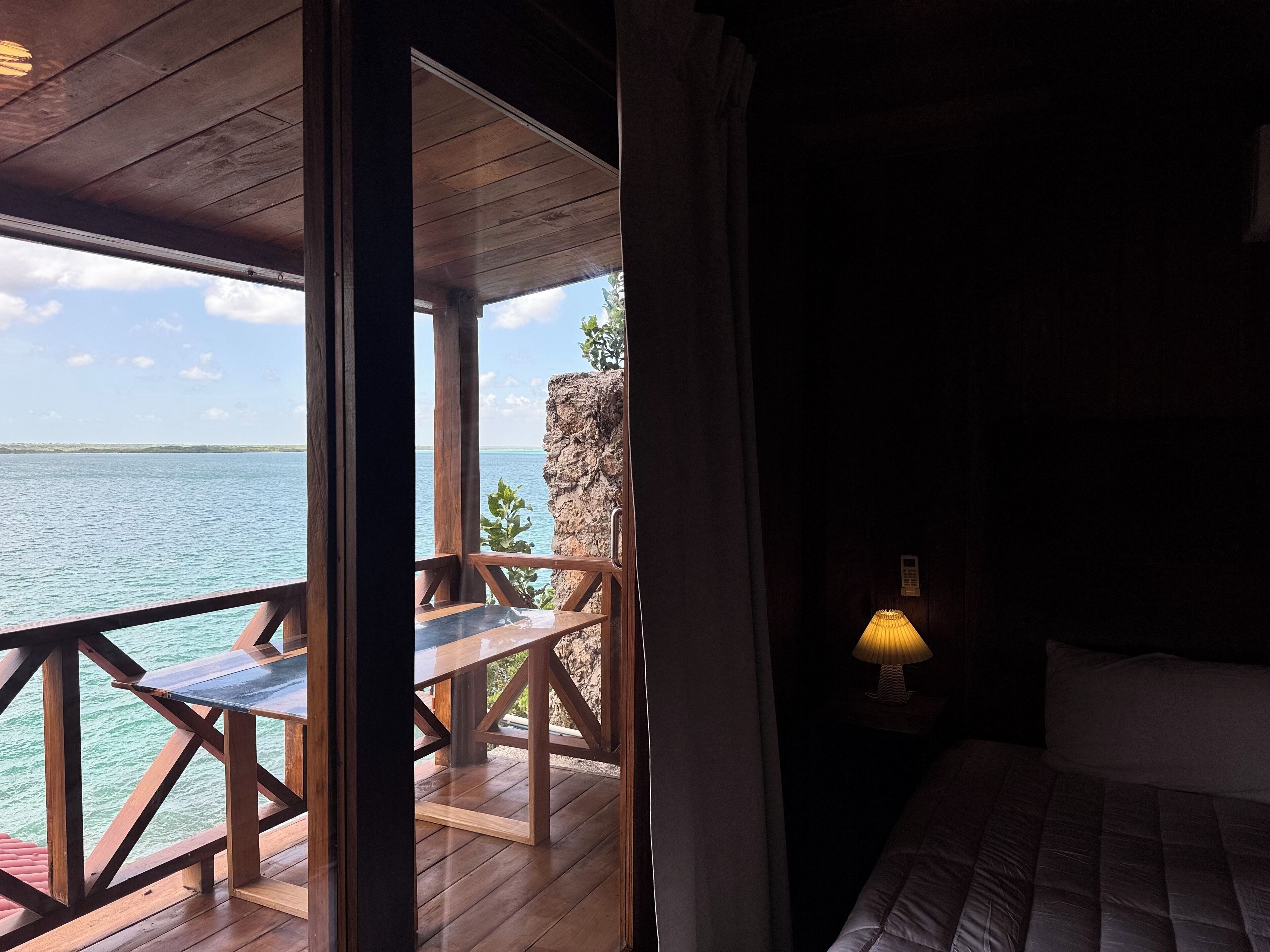 A room with a wooden deck overlooking a body of water, a bed, and a nightstand with a lamp.