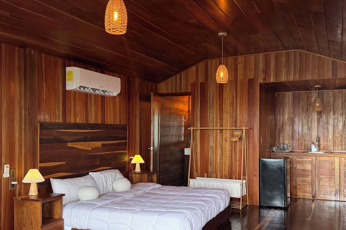 A wooden room with a bed, bedside lamps, a radiator, and a small kitchenette.