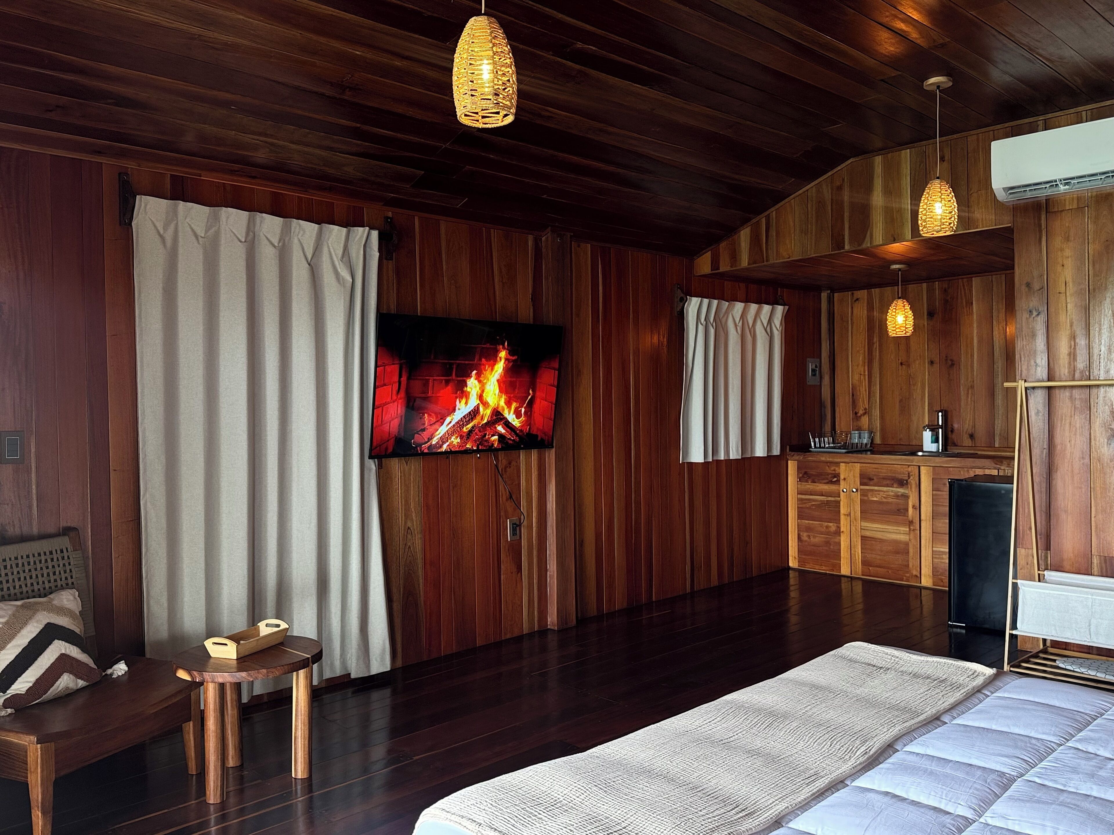 Een knus ingerichte kamer met houten details, een flatscreen televisie aan de muur, een klein houten tafeltje, een bankje en een groot bed met een witte deken.