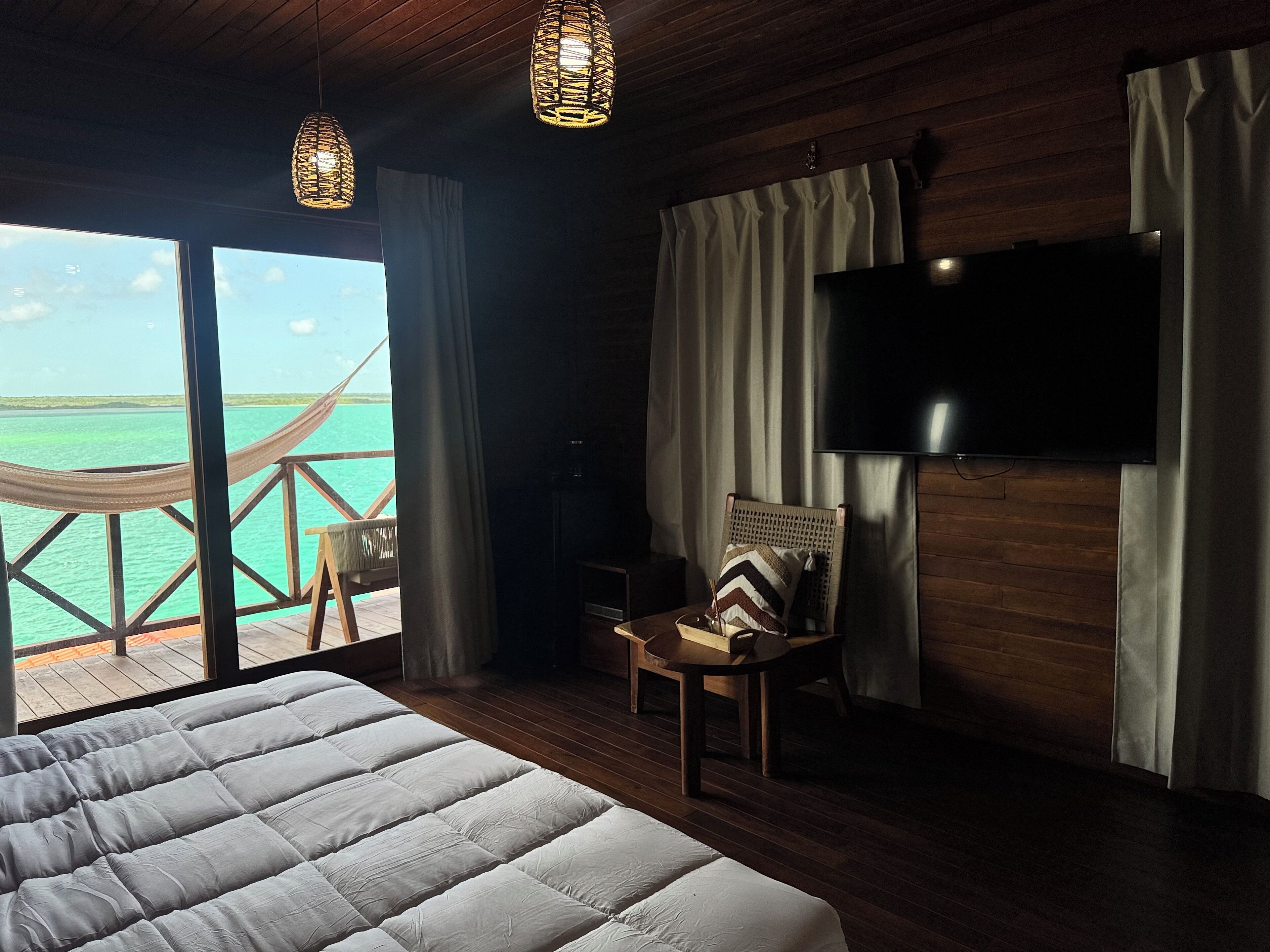 Un dormitorio con una cama grande, un televisor instalado en la pared, una silla con cojín y vistas al océano a través de una ventana.