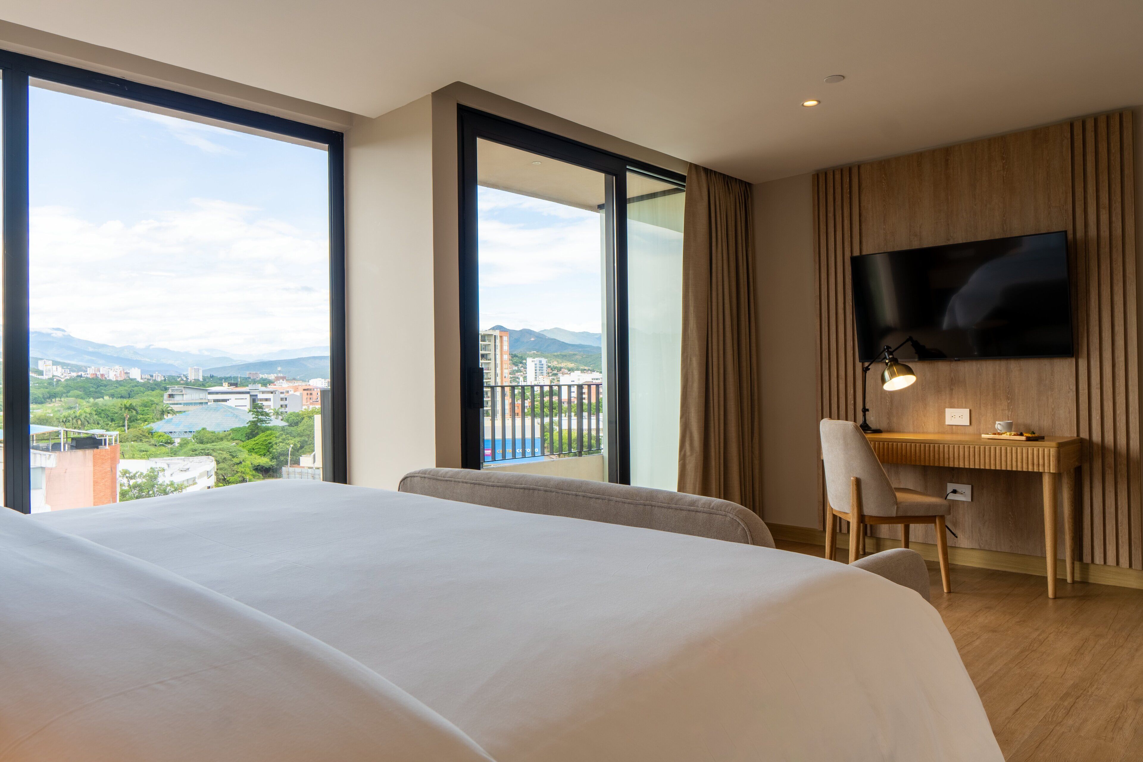 Hotelkamer met een groot bed, een bureau met stoel, een aan de muur bevestigde televisie en uitzicht op een stad en bergen door grote ramen.