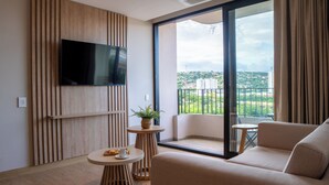 Un salon moderne avec un téléviseur à écran plat fixé sur un mur recouvert de panneaux en bois, une table ronde en bois agrémentée d’une plante, un canapé beige et un balcon offrant une vue sur la ville.