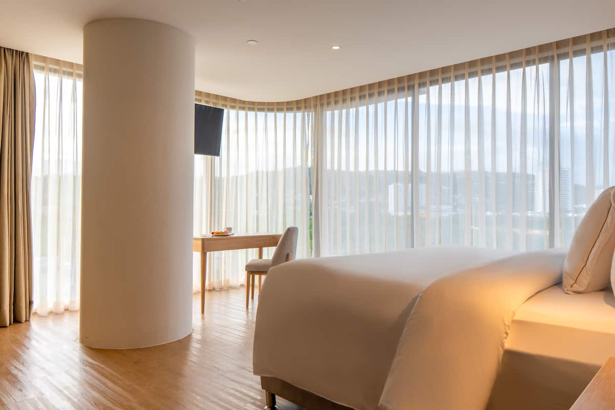 Ein Hotelzimmer mit einem großen Bett, einem Schreibtisch und einem Stuhl. An der Wand ist ein Fernseher angebracht und es gibt ein Fenster mit durchsichtigen Vorhängen.