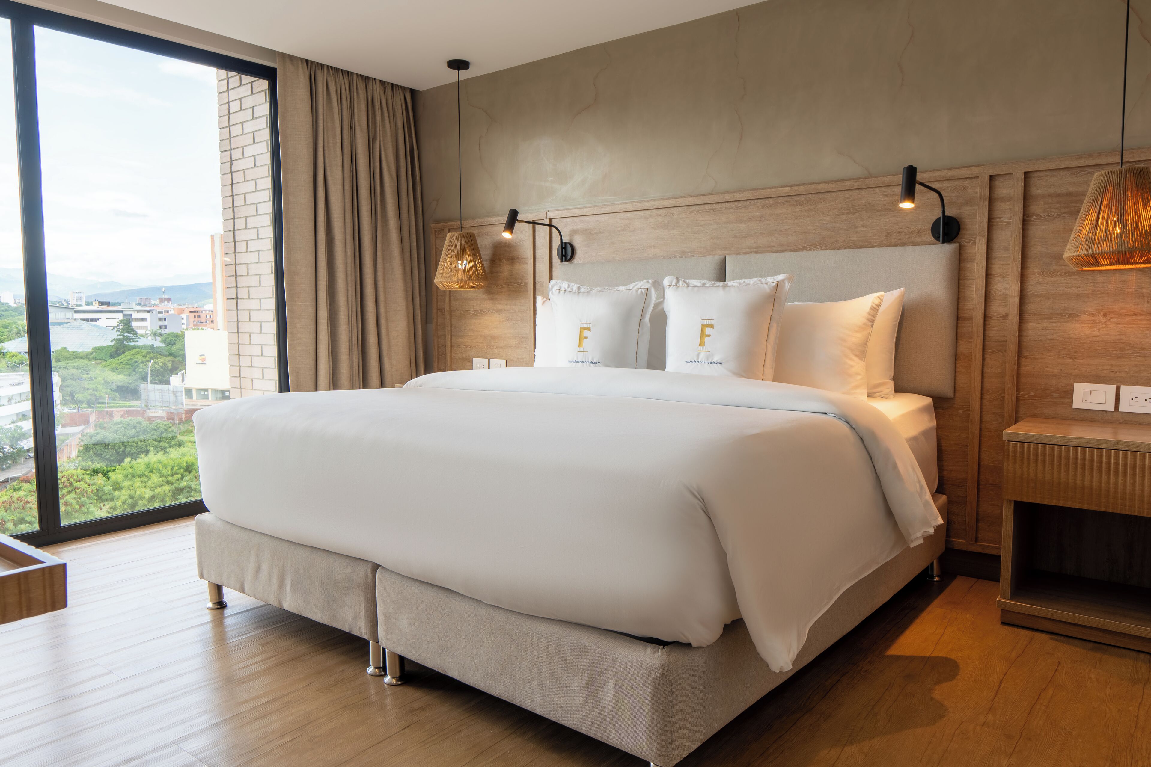 Amplia habitación de hotel con una cama grande, cabecera de madera y vistas a la ciudad a través de un ventanal.