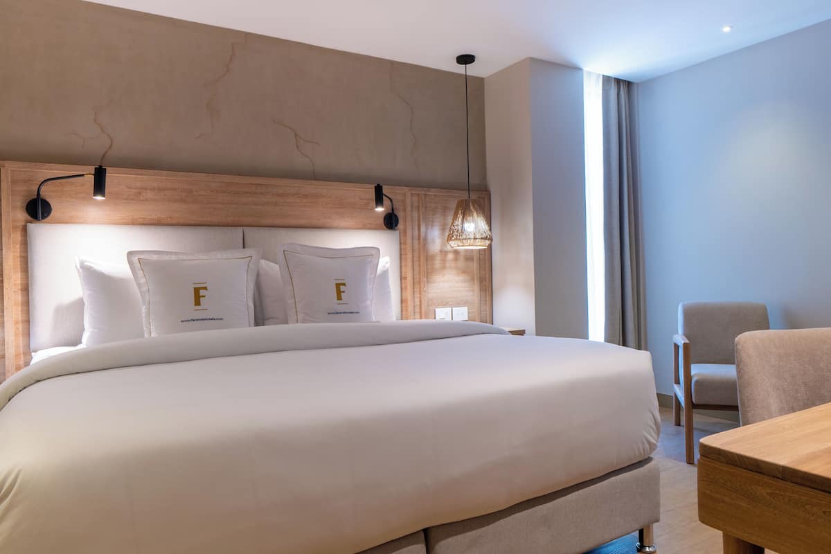 Ein modernes Hotelzimmer mit einem großen Bett, einem Holz-Kopfschild, zwei Nachttischlampen, einem kleinen Tisch und einem Sessel.