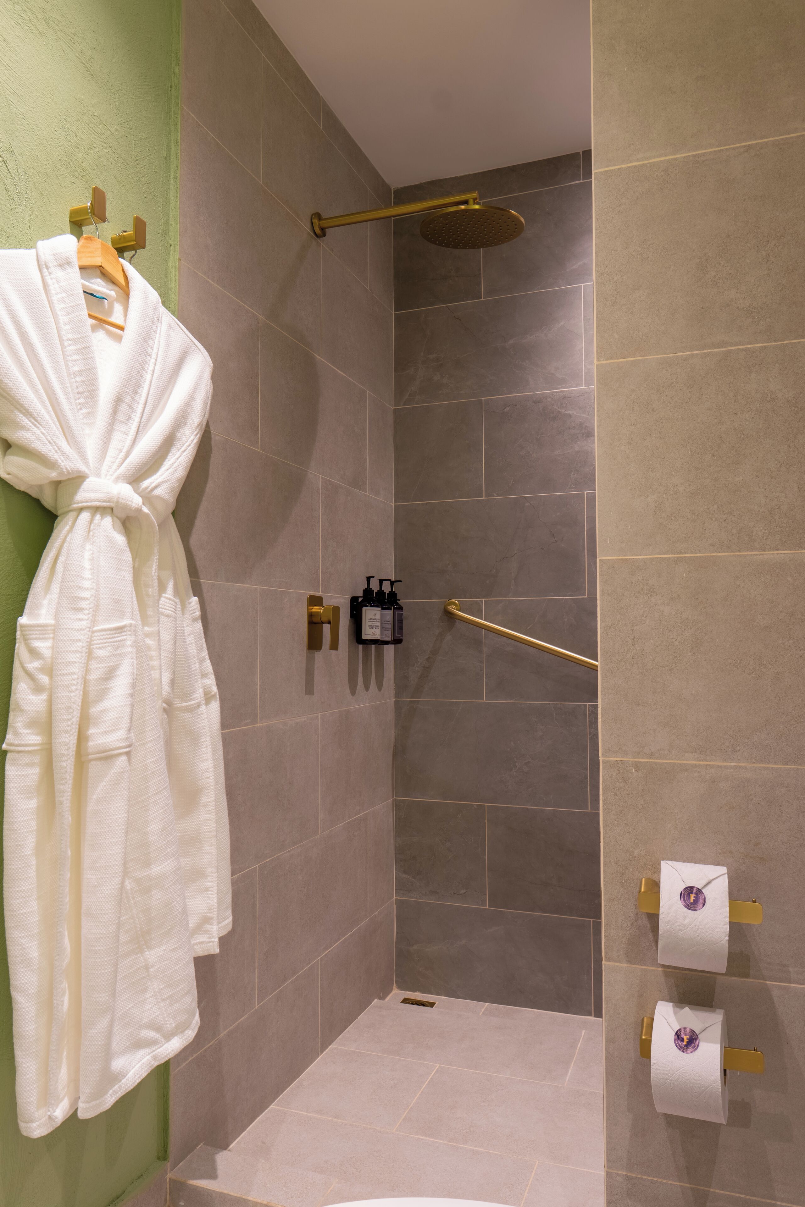 A shower area with a glass door, a towel rack with a white robe, and two bottles of shampoo and conditioner on the wall.