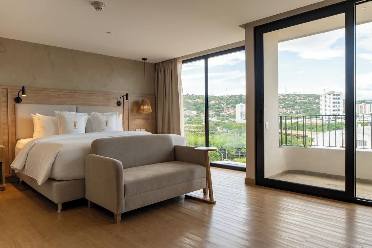 Ein modernes Hotelzimmer mit einem großen Bett, einer Sitzecke, einem kleinen Tisch und einem Balkon mit Blick auf die Stadt.