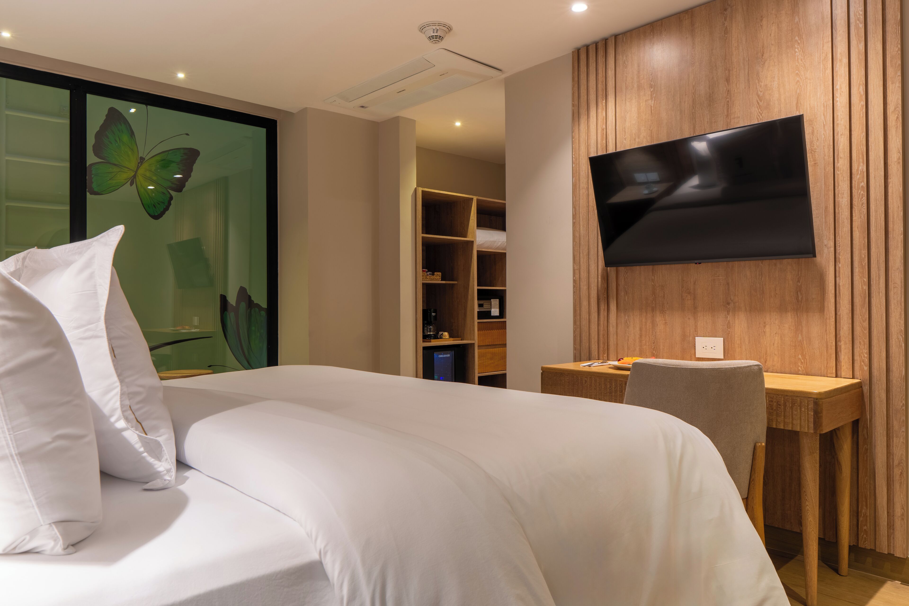 Una habitación de hotel moderna con una cama grande, un escritorio, un televisor de pantalla plana montado en la pared y un adorno de mariposa en la ventana.