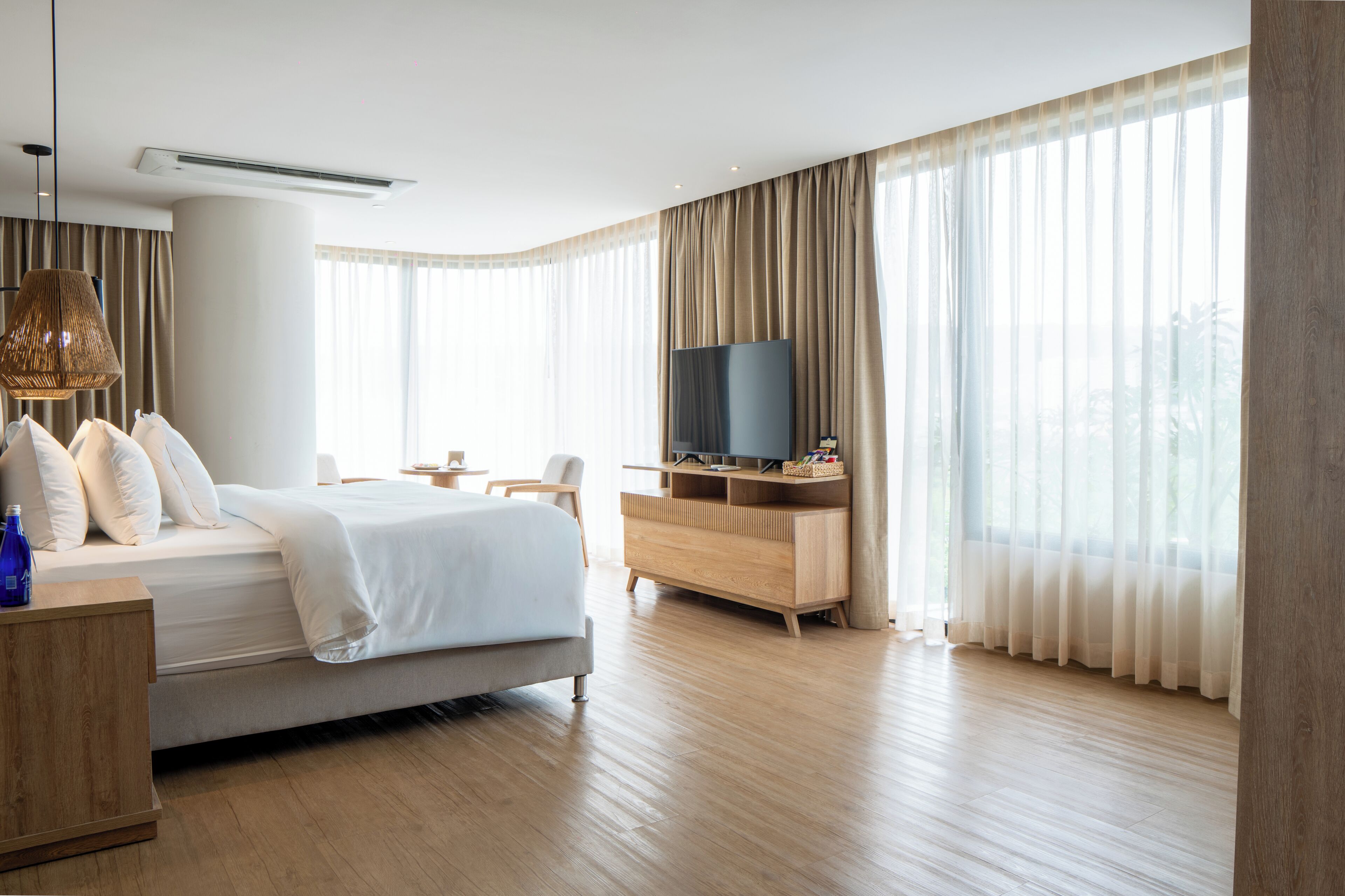 A modern hotel room with a bed, a TV, a wooden dresser, and large windows with sheer curtains.