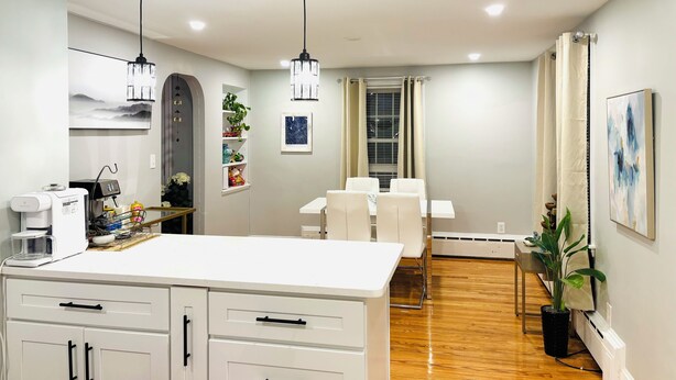 A modern kitchen with white cabinetry, a coffee machine, and pendant lighting. There is a dining area with a table and chairs, and a potted plant in the corner. - 1-bedroom unit with Private Entrance/ Laundry/Kitchen/Parking (Abington)