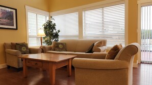 Una sala de estar con un sofá color beige, una mesa de centro de madera y una lámpara. Hay dos ventanas con persianas, una planta y un cuadro en la pared.