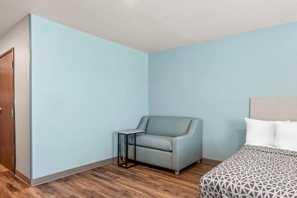 A room with a light blue wall, a wooden floor, a grey armchair, a small table, and a bed with a patterned bedspread. - Woodspring Suites Charlotte - Arrowood (Charlotte)