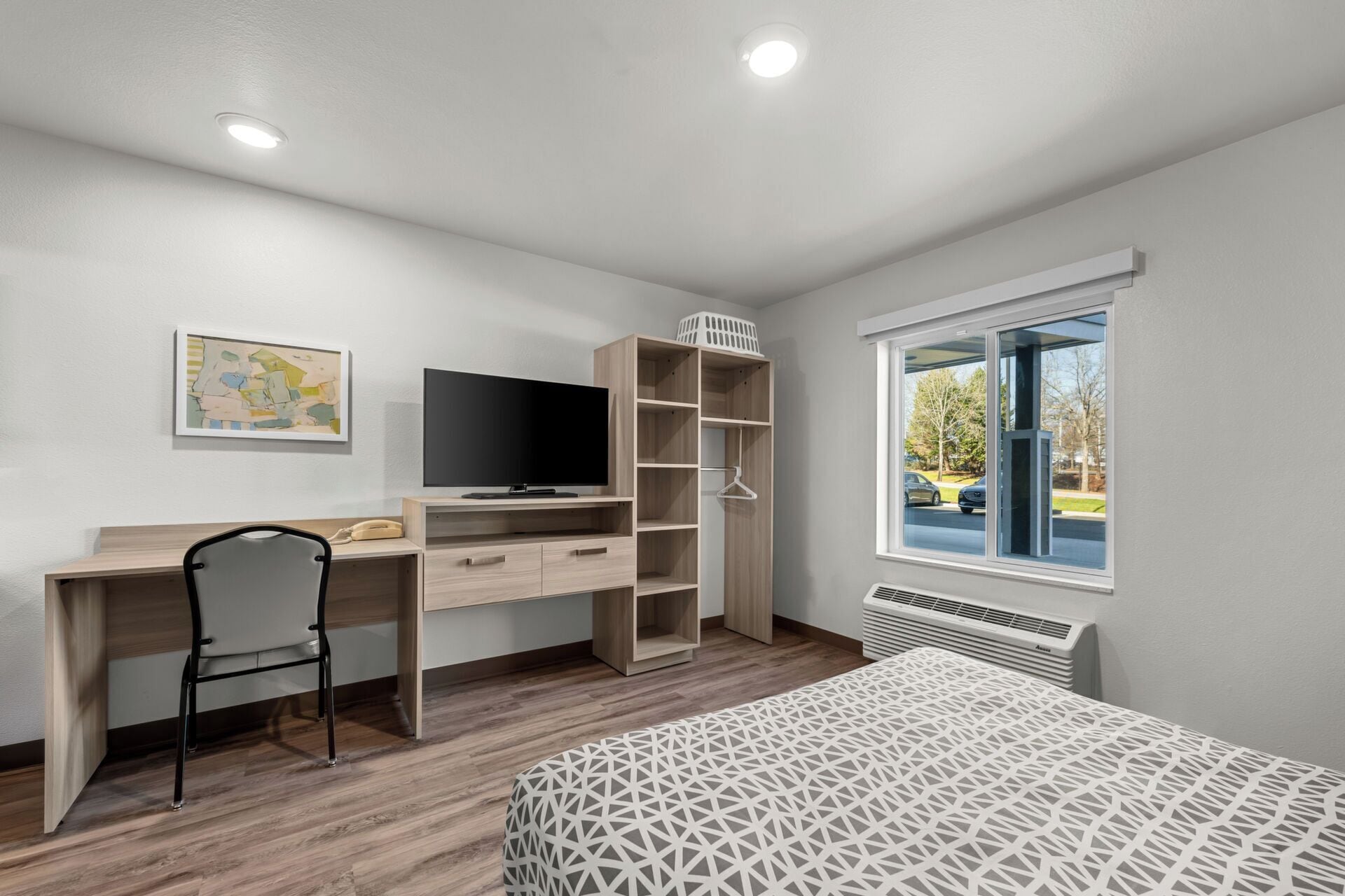 A bedroom with a wooden desk, a television, a chair, a bed with a patterned bedspread, a window with a view of the outside, and a radiator.