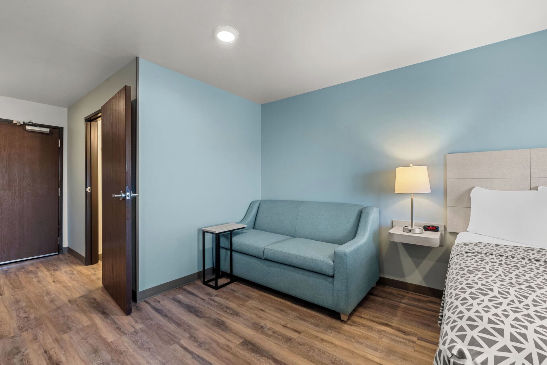 Habitación de hotel con una pared azul claro, suelo de madera, sofá, mesita de noche con lámpara y cama con colcha estampada.