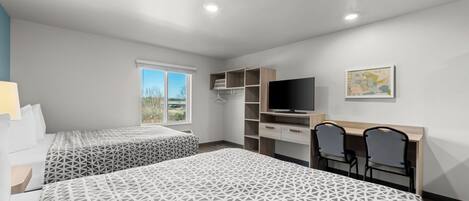 A hotel room with two beds, a TV, a desk with chairs, and a window with a view.