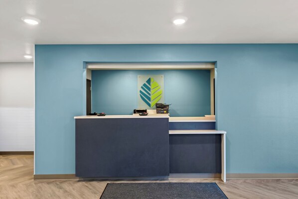 A reception desk with a blue wall, a logo on the wall, and a grey carpet. - Woodspring Suites Charlotte - Arrowood (Charlotte)