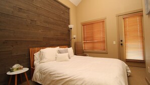 A bedroom with a wooden headboard, a bed with white bedding, a bedside table with a lamp, and a door with blinds. - Beautiful 3 Bedroom Pet Friendly Cottage on Lake Okanagan (West Kelowna)