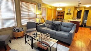 A modern living room with a gray sofa, a glass coffee table, and a fireplace. There is a dining area with a kitchen in the background. - Beautiful 3 Bedroom Pet Friendly Cottage on Lake Okanagan (West Kelowna)