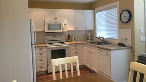 A compact kitchen with white cabinets, a microwave, stove, oven, and dishwasher. There is a window with blinds, a clock on the wall, and a bench in the center.