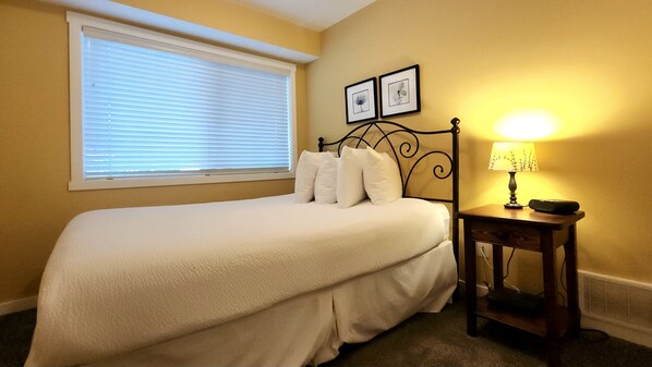A bedroom with a bed, a bedside table with a lamp, a window with blinds, and two framed pictures on the wall.
