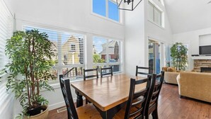 Een eetgedeelte met een houten tafel en stoelen, grote ramen voorzien van zonwering, een potplant en een bank op de achtergrond.