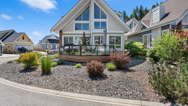 Een modern huis met twee verdiepingen, een opvallend driehoekig dak, grote ramen en een aangelegde tuin met diverse struiken en een houten terras.