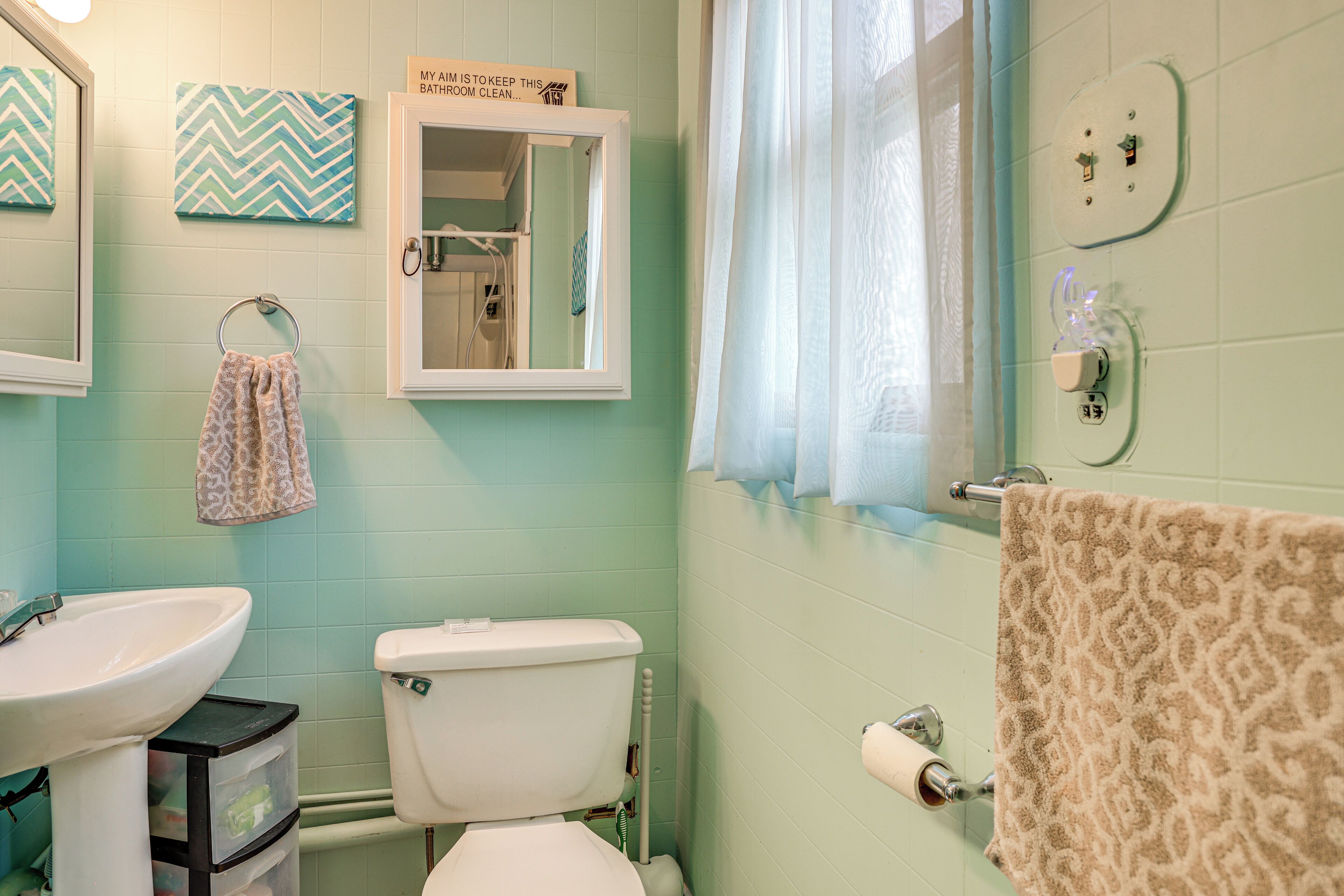 A bathroom with a sink, toilet, and a mirror. There is a towel hanging on the wall and a sign above the mirror.