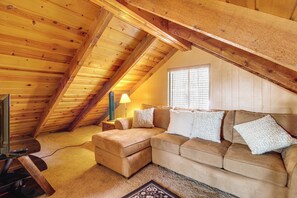 A cozy loft bedroom with a wooden ceiling, a sofa bed, a TV, and a window with blinds.
