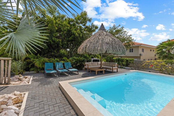 A swimming pool with lounge chairs and a thatched umbrella, surrounded by greenery and a building in the background.