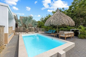 A swimming pool with lounge chairs and a thatched umbrella, surrounded by greenery and a white building.