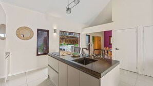A modern kitchen with a central island, a dining area with chairs, and a view of a balcony. - Villa Troupial- Jan Thiel (Jan Thiel)