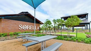 A picnic area with metal tables and benches, a brick building with "SORELLA" signage, and a covered pavilion with trees. - Patio & Pool Access: Family Home in Tomball (Hockley)