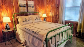 A wooden-paneled bedroom with a green metal bed frame, two bedside lamps, a pinecone lamp on a side table, and a window with blinds.