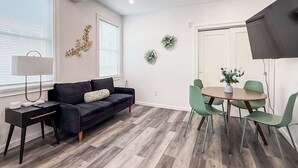 Ein modernes Wohnzimmer mit einem dunklen Sofa, einem kleinen Beistelltisch, einem runden Esstisch mit vier Stühlen und einem großen Fenster mit Jalousien.