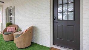 Fachada de ladrillo blanco con una puerta oscura, dos sillas de mimbre con cojines y una mesita con una planta.