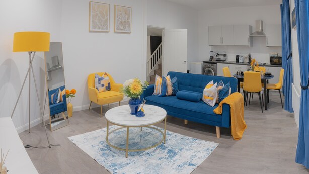 Un salon moderne avec un canapé bleu, des fauteuils jaunes, une table basse ronde et une cuisine en arrière-plan.