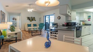 Ein modernes Wohnzimmer mit einem weißen Sofa, einem Glastisch und einem Esstisch. Im Hintergrund ist eine Küche mit weißen Schränken und einer schwarzen Arbeitsplatte zu sehen.