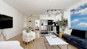 Ein modernes Wohnzimmer mit einer Couch, einem Couchtisch und einem Fernseher. Im Hintergrund ist eine Küche mit Edelstahlgeräten zu sehen.