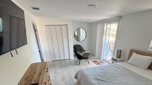 Een slaapkamer met een bed, een televisie, een houten dressoir, een stoel, een ronde spiegel en een groot raam.
