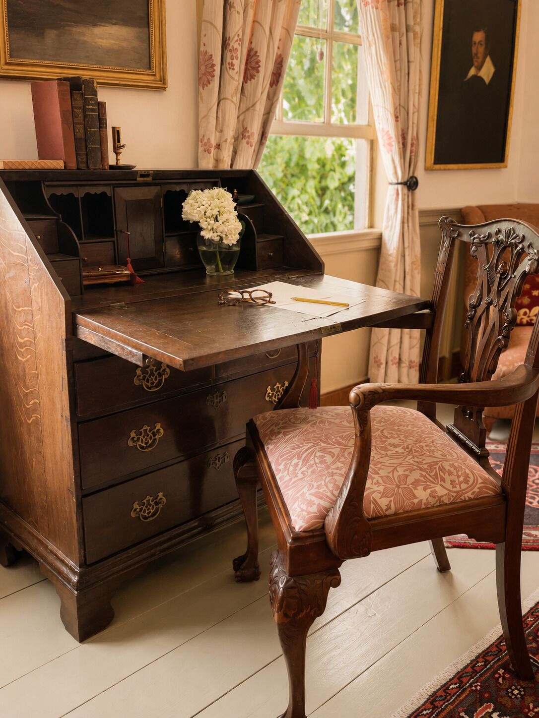 Un escritorio de madera con cajones, un jarrón con flores, una silla y una ventana con cortinas.