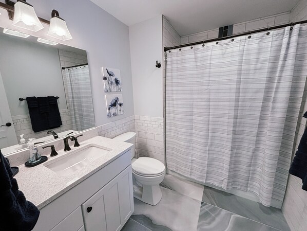 Un baño con lavamanos blanco, grifos negros, inodoro blanco, cortina de ducha y espejo con luces arriba.