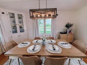 Un comedor con una mesa de madera preparada para seis personas, rodeada de sillas de mimbre, una lámpara colgante y un armario con puertas de vidrio.