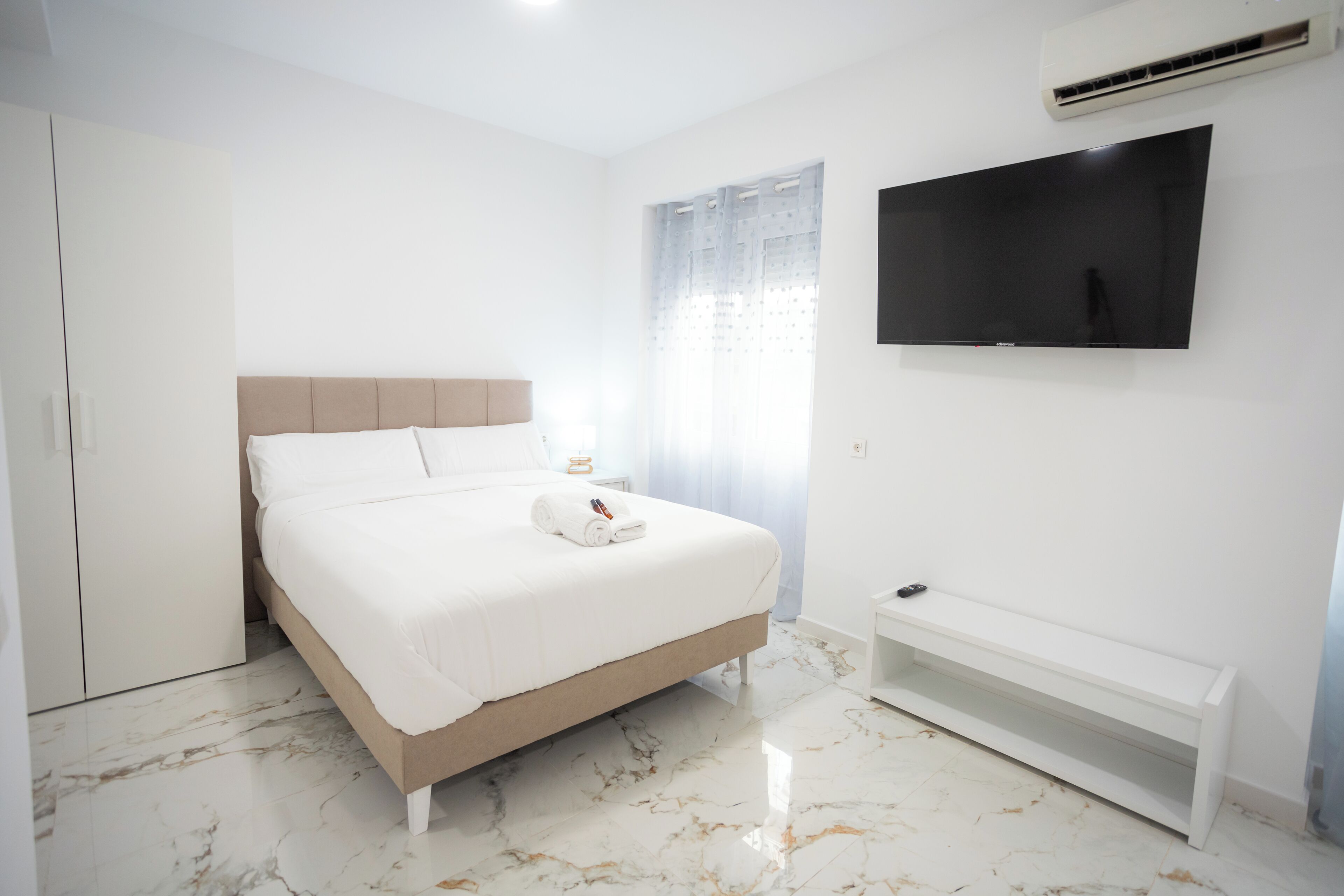 Quarto com cama, televisão na parede e um banquinho.
