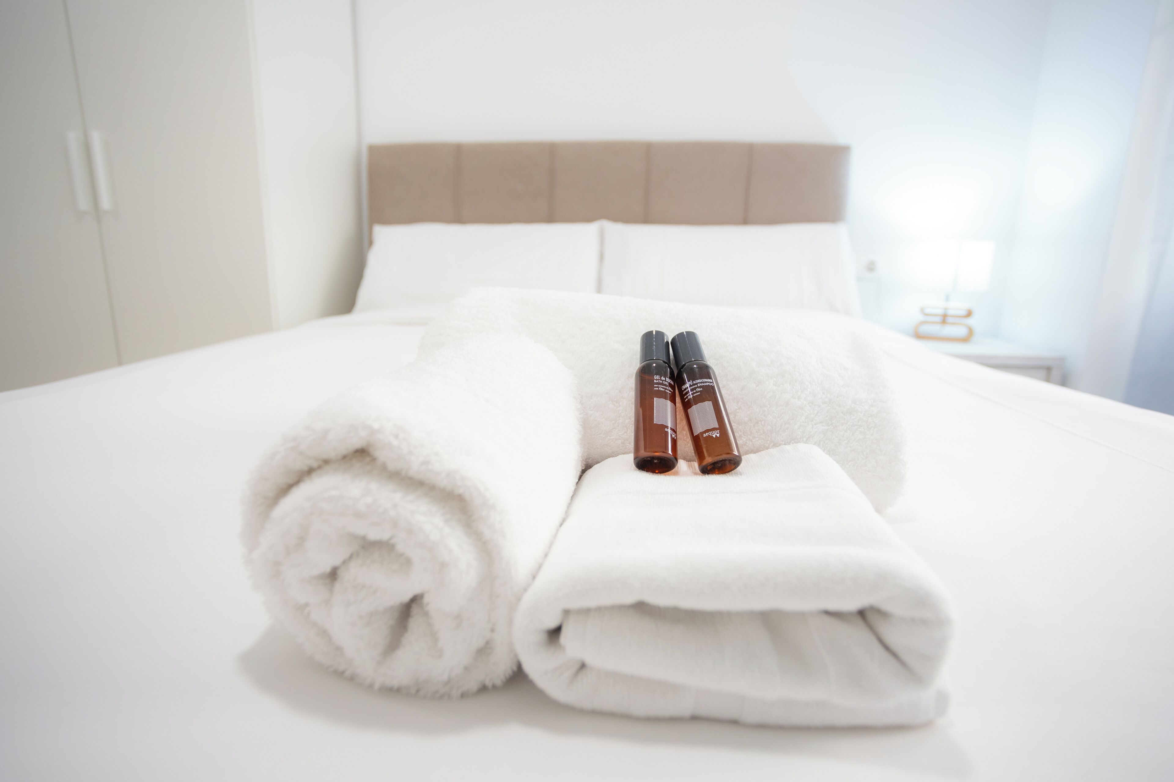 Cama bem arrumada com lençóis brancos, duas embalagens de loção e uma toalha enrolada em um quarto de hotel.