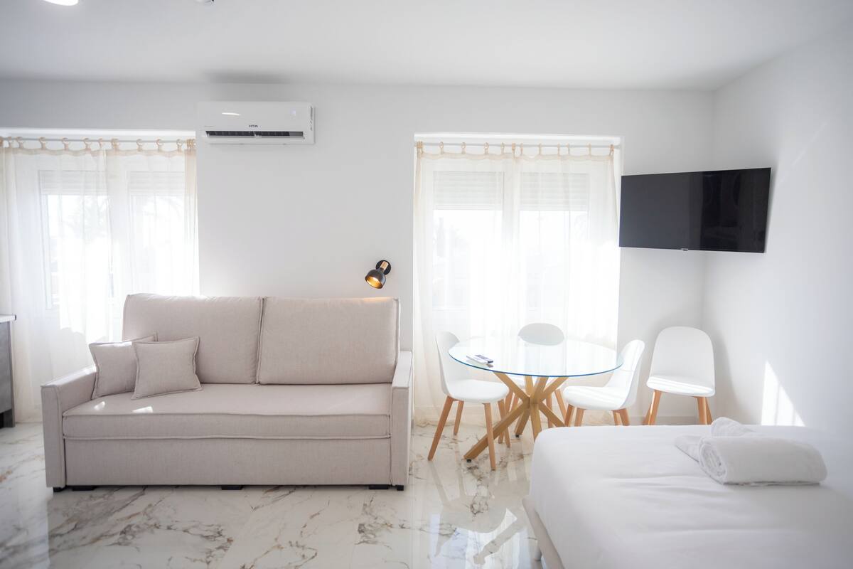Egy modern szállodai szoba, amelyben található egy kanapé, egy üveglapos asztal székekkel, egy síkképernyős televízió és egy ágy, melyen egy feltekert törölköző van.
