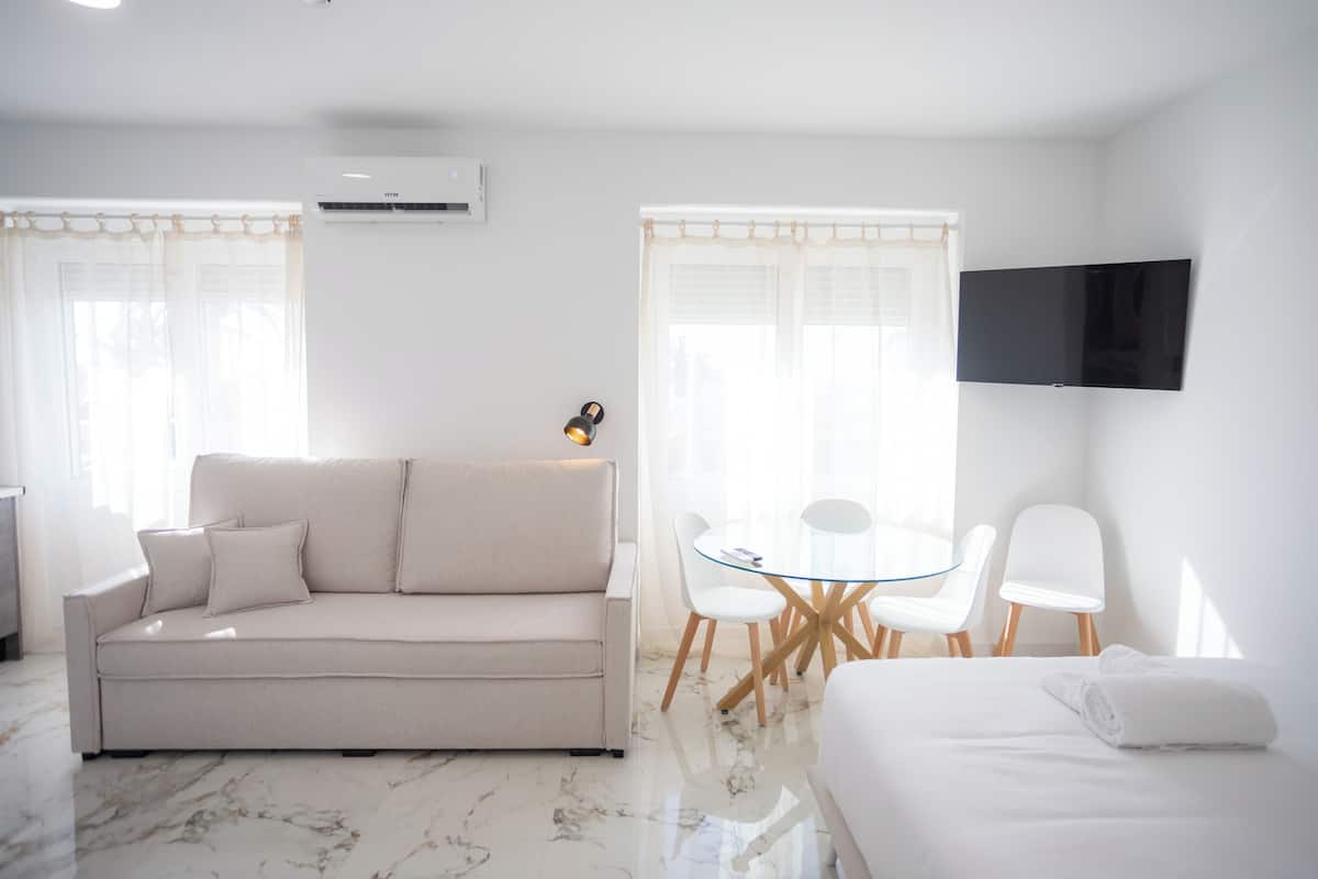 Quarto de hotel moderno com sofá, mesa de vidro com cadeiras, TV de tela plana e cama com toalha enrolada.