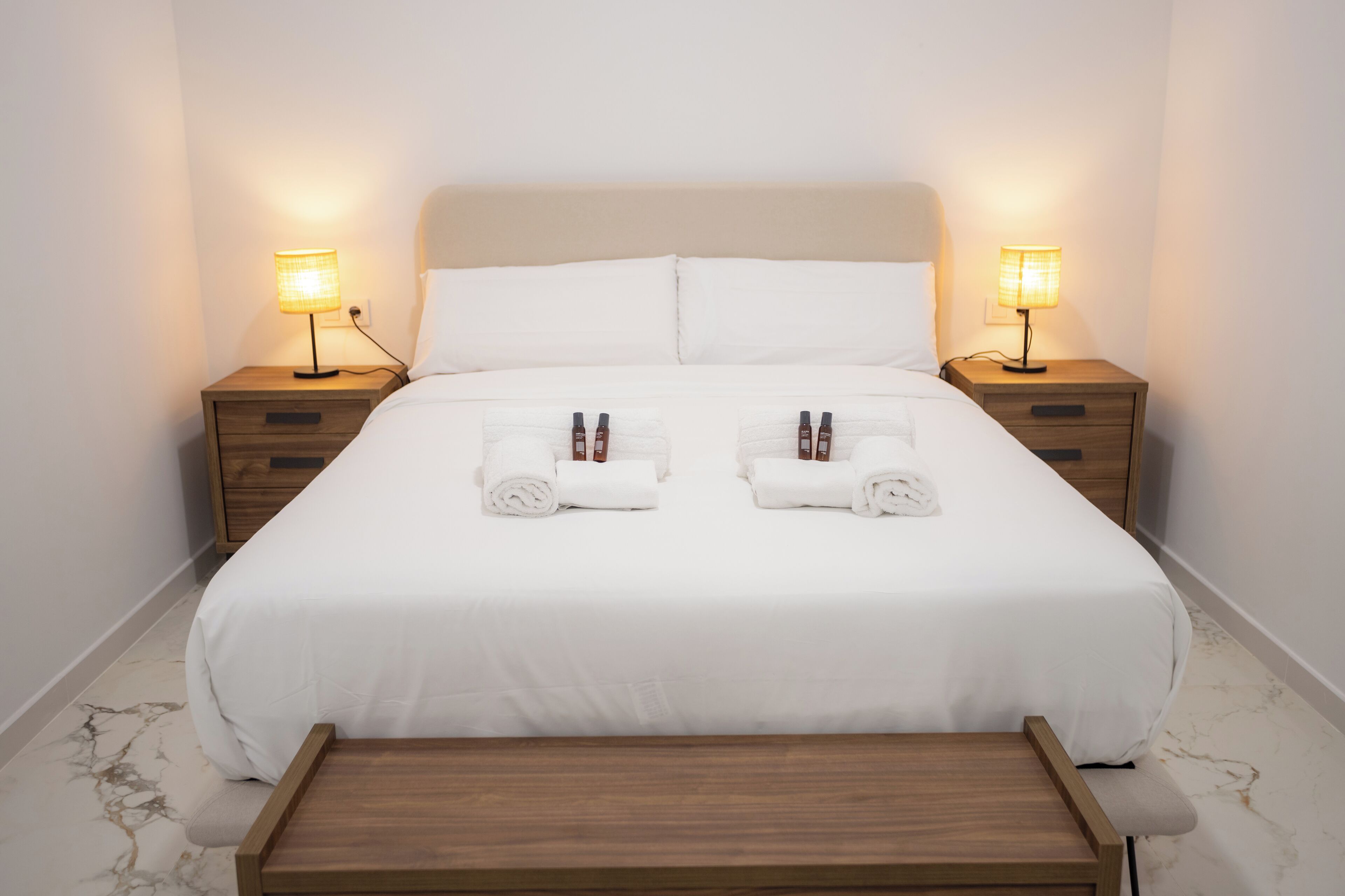 Cama bem arrumada com lençóis brancos, duas mesas de cabeceira com luminárias e um banco de madeira aos pés da cama.