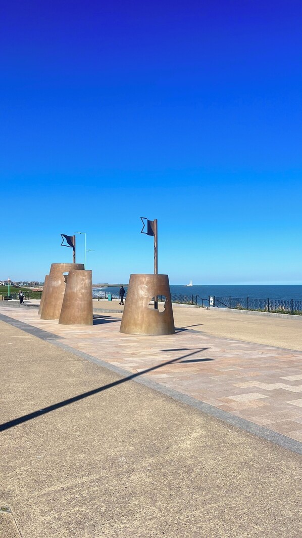 A coastal promenade with a clear sky, featuring a series of large, metallic sculptures and a distant lighthouse. - Centre of Whitley Bay Short & Long Stay (Whitley Bay)